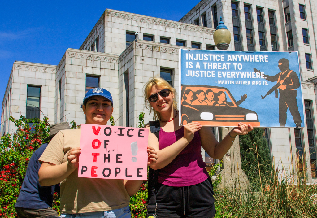 A pair of smiling women hold signs against authoritarianism and for human rights, in front of Vancouver City Hall.