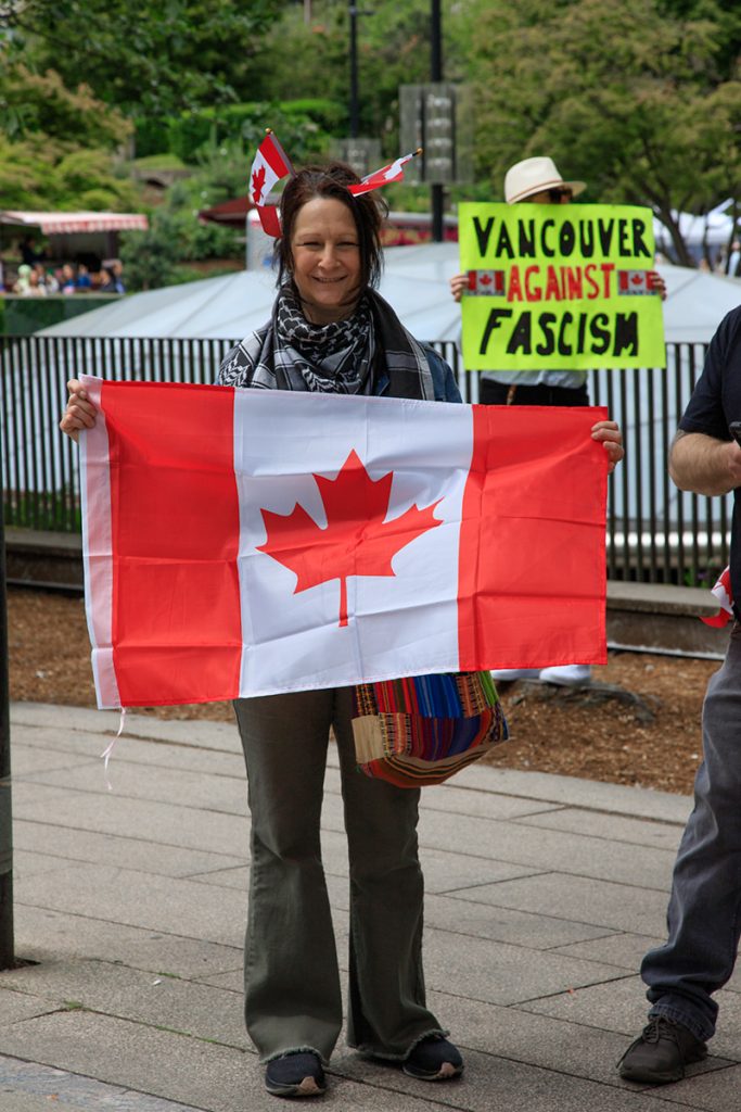 A woman holds a large Canadian flag from its top corner, and two little flags poke out of her hair like antennae. Behind her a signs reads 'Vancouver Against Fascism' - our original name.