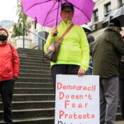 A stern woman wearing a 'Canada' baseball cap, sheltered under a pink umbrella and wearing a bright yellow coat, holding a sign which reads: 'Democracy doesn't fear protests, dictators do.'