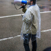 A man wearing a clear wet poncho speak into a megaphone in an street intersection.