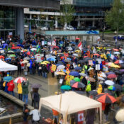 A crowd of several hundred demonstrators, sprouting with dozens of umbrellas, listing to a speaker under a small tent in Jack Poole Plaza.