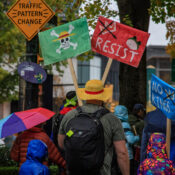 A father with a broad back, wet in the rain, with his two kids from behind, holding signs saying Resist and No Kings and a street sign reading "Traffic Pattern Change"