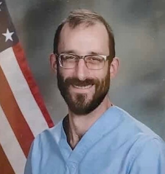 Portrait of Alex Pretti, in his medical gown, smiling, warm, bearded with glass. He was murdered today by ICE in Minneapolis.