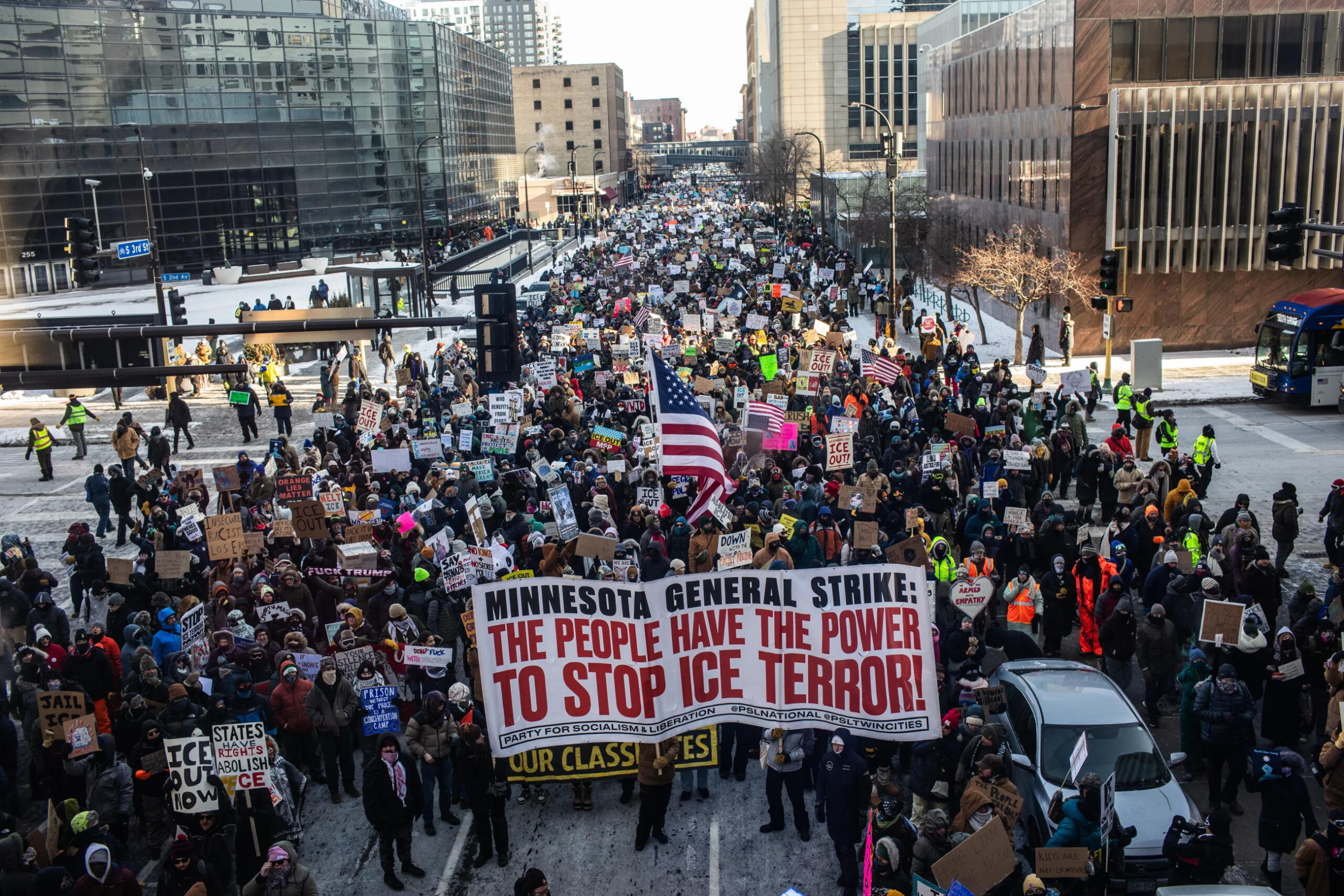 From the air, a packed crowd filling a six lane city road into the far distance, wearing winter clothing and carrying anti-ice signs, including one that reads "Minnesota General Strike: the people have the power to stop ice terror
