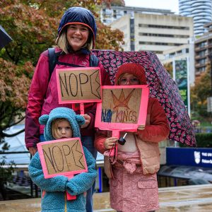 nope-nope_jdowler_3641_900x900px A smiling mother and her two shy kids dressed in pink holding to signs reading 'Nope' and one saying 'No Kings'